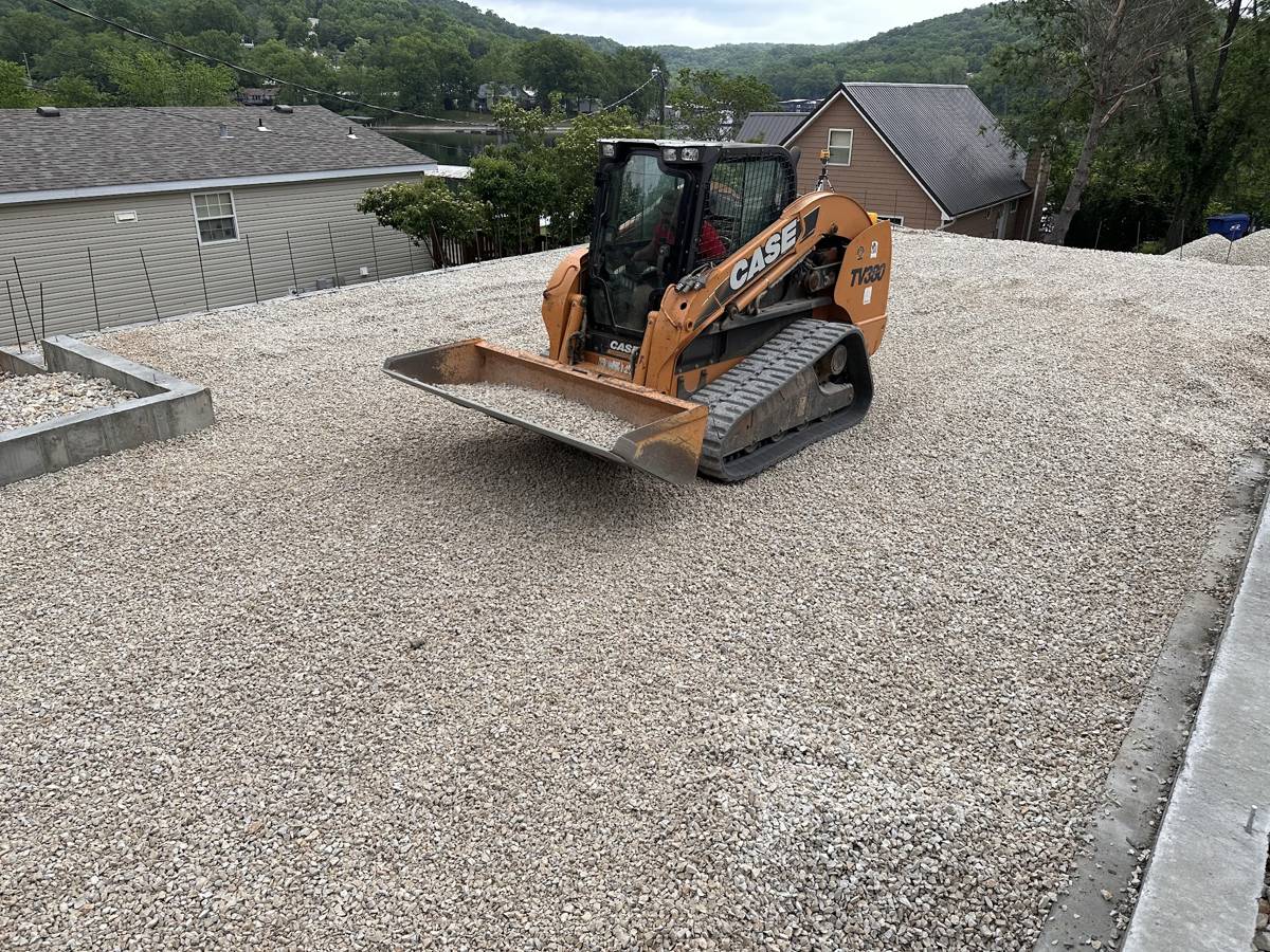 Track loader spreading fresh gravel on a sloped lot with lake hills behind